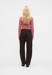 Femme blonde portant un haut rose à manches longues, un pantalon large foncé avec poches arrière et des chaussures à talons noires, debout de dos sur fond blanc.