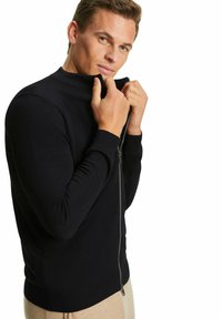 Schwarzer Reißverschluss-Pullover mit hohem Kragen, gerippten Bündchen und glatter Textur. Das Modell zieht mit beiden Händen am Reißverschluss.