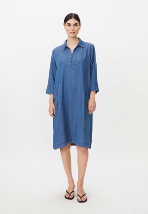 Kvinde med mørkt hår iført en løs blå denimkjole med trekvart ærmer og sorte sandaler, der står mod en ensfarvet lys baggrund.