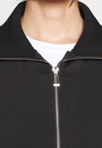 Giacca nera con zip in lurex, caratterizzata da un colletto a costine e una cerniera argentata, che mette in mostra una superficie liscia e un design minimalista.