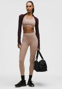 Sportoutfit mit einem ziegelroten Langarmshirt, beigem BH und beigen high-waist Leggings; kombiniert mit schwarzen Turnschuhen und einer schwarzen Sporttasche.