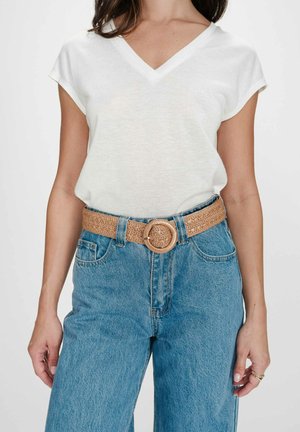 Femme portant un haut blanc à manches courtes en col V, rentré dans un jean bleu taille haute avec une ceinture tressée couleur camel à boucle ronde.