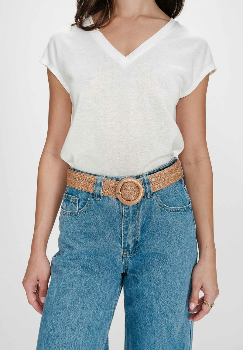 Femme portant un haut blanc à manches courtes en col V, rentré dans un jean bleu taille haute avec une ceinture tressée couleur camel à boucle ronde.