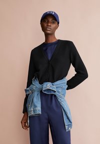 Cardigan nero con scollo a V e bottoni, indossato sopra un top blu navy, con una giacca di denim legata in vita. Texture morbida; vestibilità ampia.