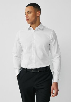 Mann in weißem Hemd mit Knöpfen und schwarzer Hose, mit teilweise in den Taschen steckenden Händen, der seitlich gegen einen schlichten grauen Hintergrund blickt.