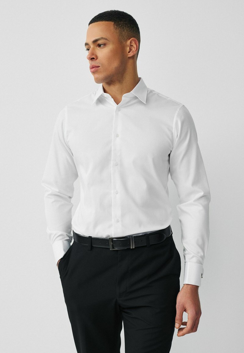 Homme portant une chemise blanche boutonnée et un pantalon noir, les mains partiellement dans les poches, regardant sur le côté sur un fond gris uni.