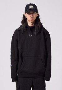 Black hoodie with a kangaroo pocket, drawstrings, and graphic detail on the left sleeve. Paired with a black cap featuring an embroidered logo.