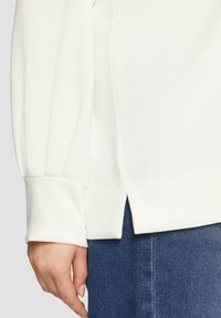 Witte lange mouwtop met een afgeronde onderkant en zijslitsen, gemaakt van gladde stof, gecombineerd met donkerblauwe denim. Close-up van de mouw en hand.