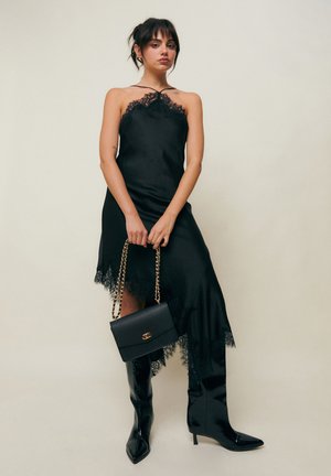 Žena v čiernych saténových šatách s čipkovaným lemom, drží čiernu kabelku so zlatými retiazkovými popruhmi, v kombinácii s čiernymi čižmami nad kolená so špičatou špičkou.