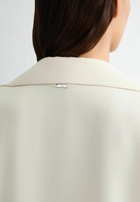Světle béžová blůza s hladkou texturou, s límečkem a stříbrnou logovkou na zadní straně. Jednoduchý a elegantní design.
