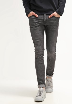 Personne portant un jean slim gris foncé avec les revers retroussés, les mains dans les poches, et des baskets gris clair marchant vers l'avant.