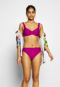 Magentan värinen bikiniyläosa, jossa on tukirauta ja leveät olkaimet, yhdistetty korkeavyötäröisiin magentan värisiin alaosiin. Malli käyttää kukkakuosista päällystakkia.