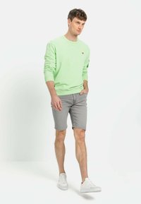 Helltgrün sweatshirt mit rundem Ausschnitt, grauem Denim-Shorts und weißen Sneakers. Das Sweatshirt hat ein kleines Emblem auf der Brust.