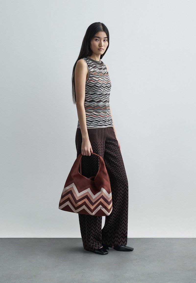 Une femme se tient debout, tenant un sac à main marron à motifs zigzag, portant un haut sans manches à motifs et un pantalon large à rayures zigzag assorti de couleur sombre.