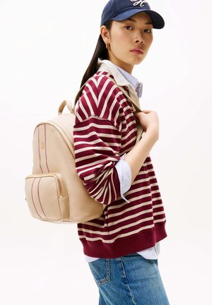 Giovane persona che indossa un maglione a righe color bordeaux, un berretto blu e jeans, con uno zaino beige su una spalla, di profilo.