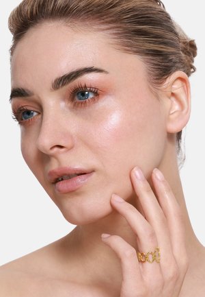 Goldring mit verknüpften kreisförmigen Elementen, der an einem Finger getragen wird; die Haut erscheint glatt mit einem leichten Glanz; dezentes Makeup auf dem Gesicht.