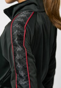 Schwarze Jacke mit höherem Kragen, versehen mit einem glänzenden schwarzen Logomuster entlang des Ärmels und roten Akzentpaspelierungen. Hergestellt aus strukturiertem Stoff.