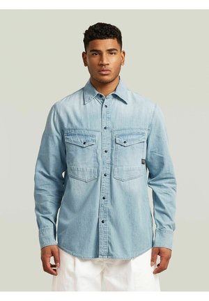 Man met kort krullend haar, die een lichtblauw denim overhemd met knopen en borstzakken draagt en een witte broek, staand tegen een effen achtergrond.
