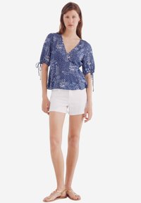 Blusa azzurra con motivo e maniche puff corte, scollatura a V, abbinata a pantaloni corti bianchi e sandali piatti dorati.