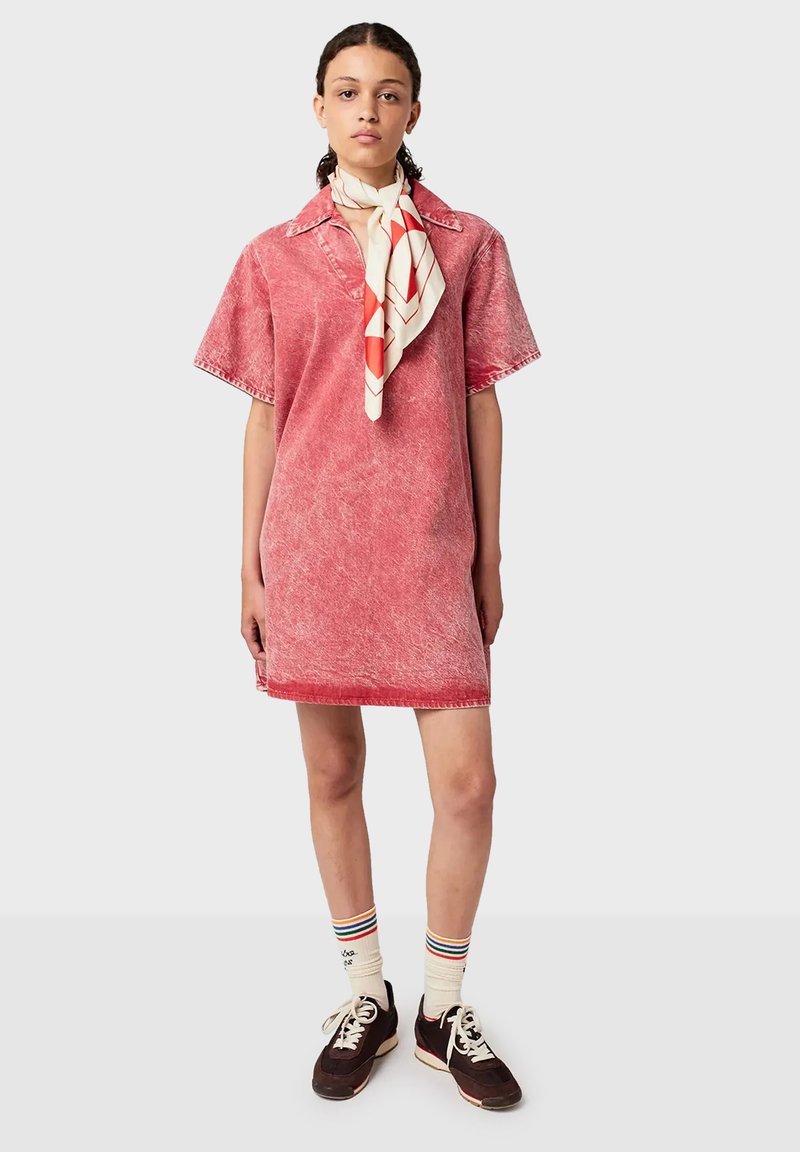 Jonge vrouw in een vervaagd rode jurk met korte mouwen, een crèmekleurige en rode gedessineerde sjaal om haar nek, gestreepte sokken en donkere sneakers tegen een neutrale achtergrond.
