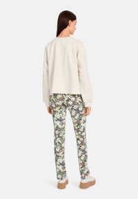 Cremfarbener Pullover mit langen Ärmeln, kombiniert mit gemusterten Hosen mit floralen und grafischen Designs in Grün, Gelb und Braun. Weiße Sneakers.