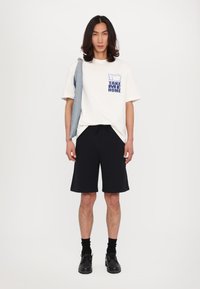 Witte katoenen t-shirt met blauwe grafische opdruk "TAKE ME HOME" op de borst, gecombineerd met zwarte short. Man houdt een spijkerjack over zijn schouder. Zwarte schoenen.