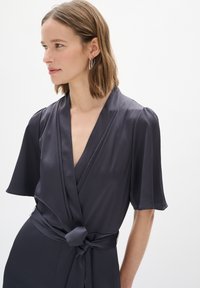 Abito avvolgente grigio scuro con maniche corte, tessuto strutturato, scollatura profonda a V e dettaglio in vita con cintura, completato da minimi accessori metallici.