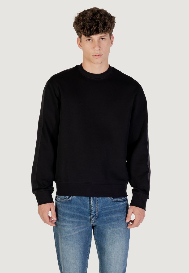 Joven con pelo rizado, vistiendo un sudadera negra básica y jeans azules, de pie contra un fondo gris claro.