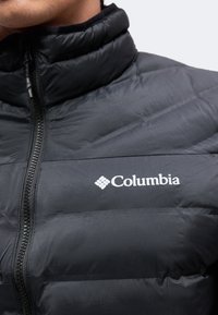 Gros plan d’une personne portant une doudoune noire avec une fermeture éclair devant et un logo Columbia blanc sur la poitrine.