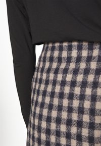 Gros plan sur une personne portant un haut à manches longues noir rentré dans une jupe taille haute à carreaux noirs et beige.