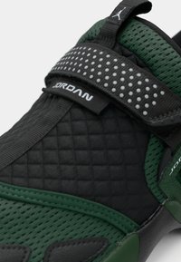 Zwarte en groene sneaker met een gestructureerde bovenkant, gevoerde kraag, verstelbare riem en geperforeerde accenten. Bevat "JORDAN" branding.