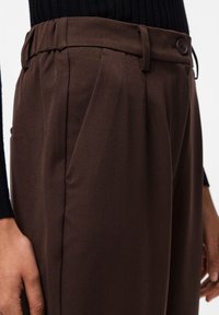 Braune maßgeschneiderte Hose aus einem strukturierten Stoff, mit einem Bund mit einem Knopf und Falten, seitlich präsentiert.