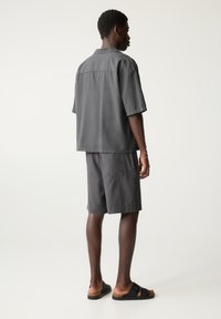 Grå kortærmet skjorte og shorts sæt, lavet af blødt stof. Shortsene har en sidelomme. Sorte slidesandaler fuldender looket.