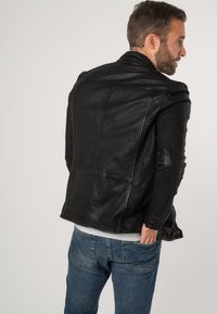 Giacca di pelle nera con finitura texturizzata, caratterizzata da una vestibilità aderente e cuciture strutturate, abbinata a jeans blu chiaro.