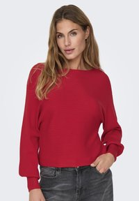 Red ribbed sweater with a wide neckline and long, slightly puffed sleeves. Worn over dark gray jeans, featuring a relaxed fit and smooth texture.