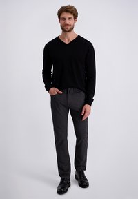 Zwarte lange mouwen V-hals trui gecombineerd met grijze getextureerde broek en zwarte veterschoenen. Model staat met handen in de zakken tegen een grijze achtergrond.