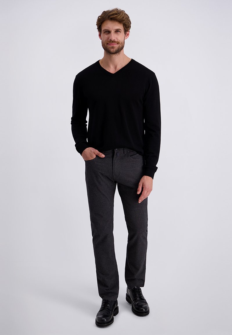 Zwarte lange mouwen V-hals trui gecombineerd met grijze getextureerde broek en zwarte veterschoenen. Model staat met handen in de zakken tegen een grijze achtergrond.