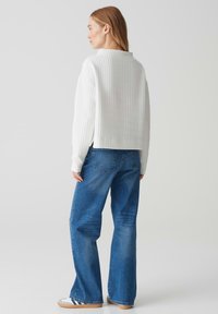 Weißes, geripptes Langarmoberteil mit kurzem Schnitt. Getragen mit blauen, geraden Jeans und weißen Sneakers mit blauen Akzenten. Minimalistischer Stil.