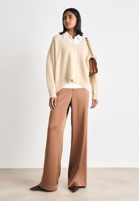 Beige oversized sweater with V-neck, paired with loose brown satin trousers and pointed brown heels. Woman holds a brown crossbody bag.