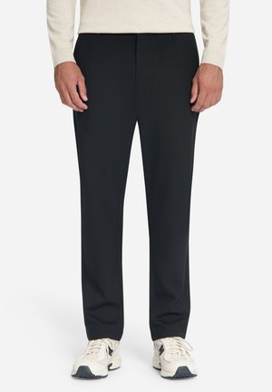 Man draagt zwarte rechte broek, beige trui en witte sneakers, staat met ontspannen handen langs het lichaam.