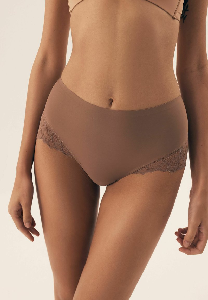 Kobieta w jasnobrownym, bezszwowym bieliźnie z koronkowym wykończeniem, pokazana od talii do połowy uda, stojąca na jednolitym tle.