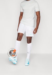 Světle šedé sportovní tričko Nike s tyrkysovými akcenty, bílé pruhované šortky, bílé ponožky a světle modré kopačky, držící černobílý fotbalový míč.