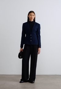 Μπλε σκούρο σακάκι με αιχμηλές πέτσες, δύο ασημένια κουμπιά, συνδυασμένο με μαύρο παντελόνι φαρδύ και μικρή μαύρη τσάντα χειρός.