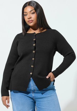 Ung kvinde med glat mørkt hår iført en sort knap-op cardigan og lyseblå jeans, stående med den ene hånd i lommen.