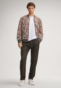 Kurtka bomber z beżowego wzorzystego materiału z czerwonymi ustami, szarymi ściągaczami, biała koszulka, ciemnozielone spodnie i białe sneakersy.