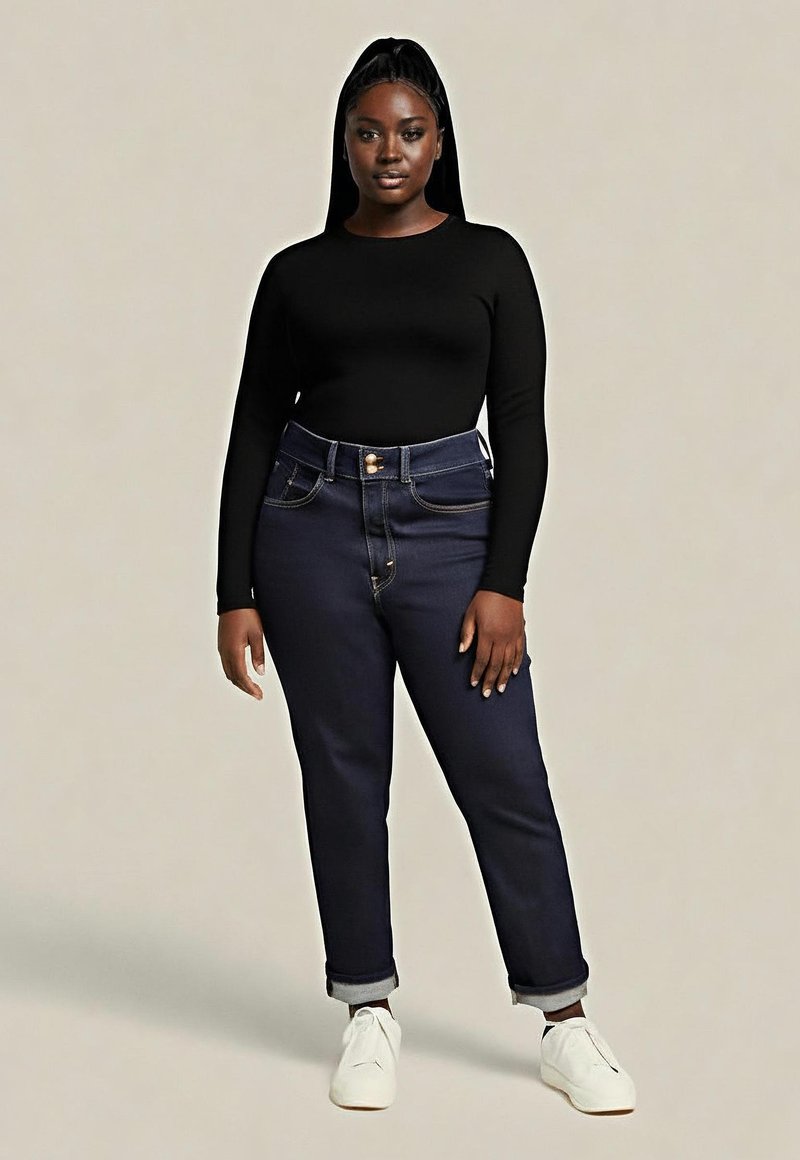 Jeans en denim foncé à taille haute, avec des ourlets retroussés et des coutures jaunes contrastantes, associés à un haut noir à manches longues et des baskets blanches.