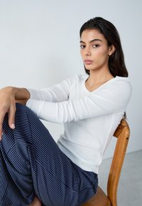 Top bianco a maniche lunghe a costine abbinato a pantaloni a gamba larga blu scuro e bianchi a righe. Il modello è seduto su una sedia di legno contro uno sfondo chiaro.