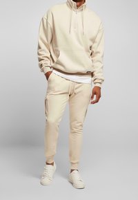 Uomo che indossa un hoodie beige a mezza zip, pantaloni jogger abbinati con tasche laterali, una maglietta bianca sotto e sneakers bianche su uno sfondo neutro.
