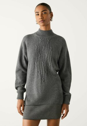 Kvinde iført en grå strikket sweater med kabelmønstre og matchende grå nederdel, stående mod en ensfarvet hvid baggrund.