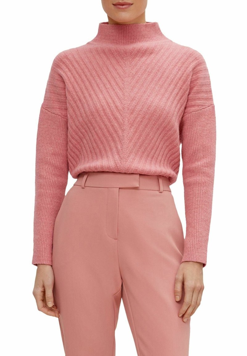 Femme portant un pull à col roulé côtelé rose et un pantalon taille haute assorti, debout, les bras détendus le long du corps.